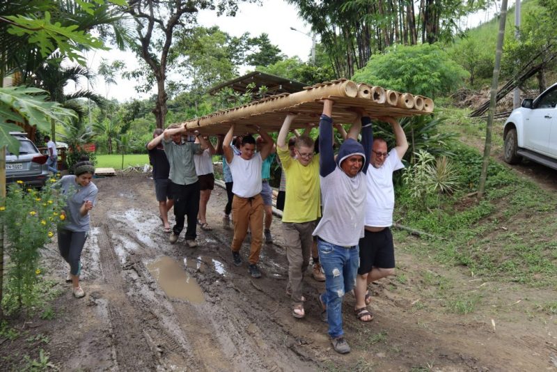 A group carries a plank built of assembled bamboo above their heads down a muddy path